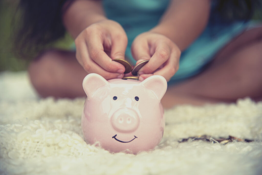 Dłonie dziecka wkładającego monety do różowej, uśmiechniętej świnki skarbonki.