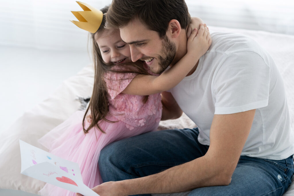 Młody tata przytula kilkuletnią córkę w stroju księżniczki, która daje mu laurkę