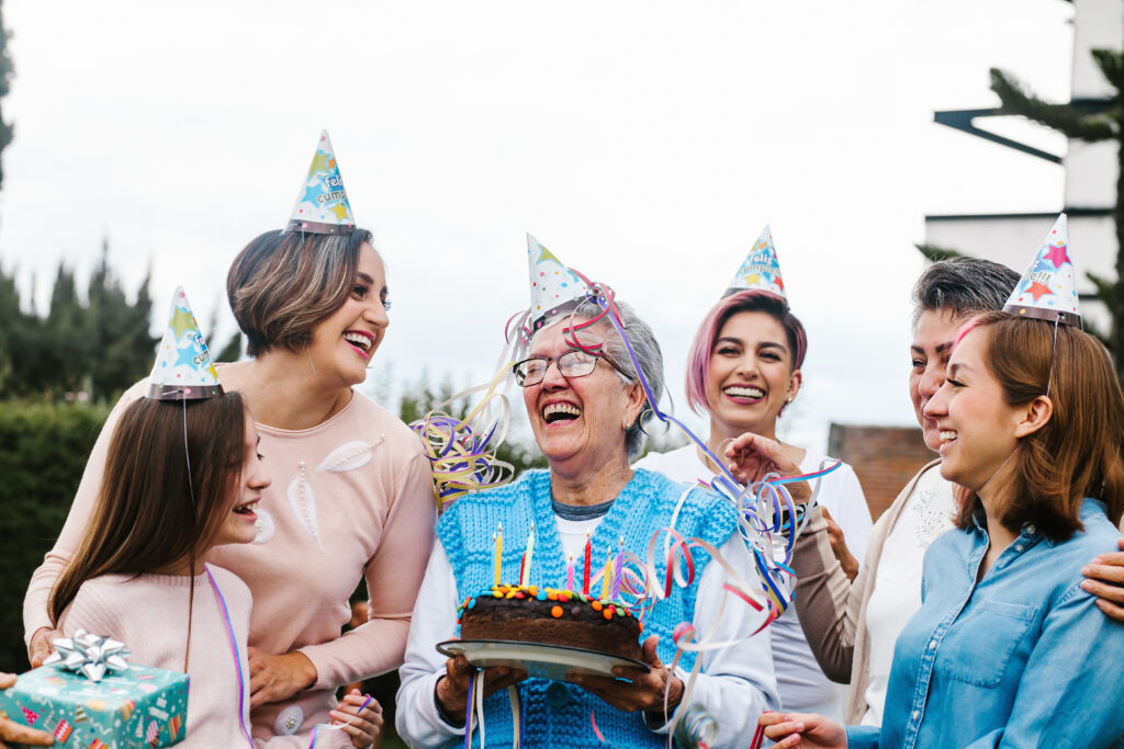 Seniorka trzyma w dłoniach tort urodzinowy i świętuje z rodziną
