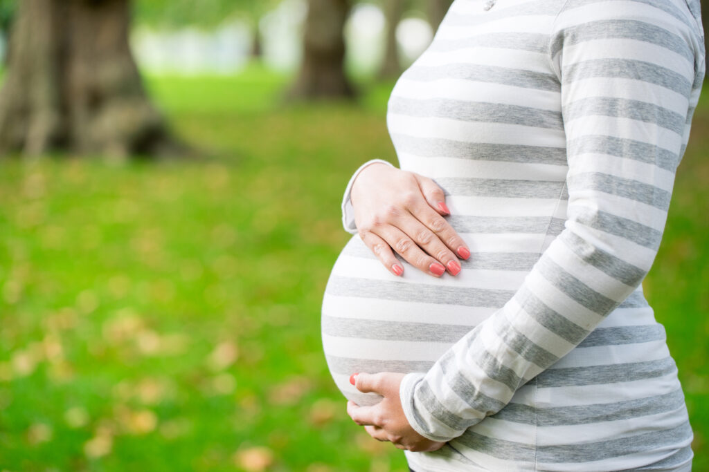 Sylwetka kobiety w ciąży, w bluzce w szaro-białe pasy, na tle parku.